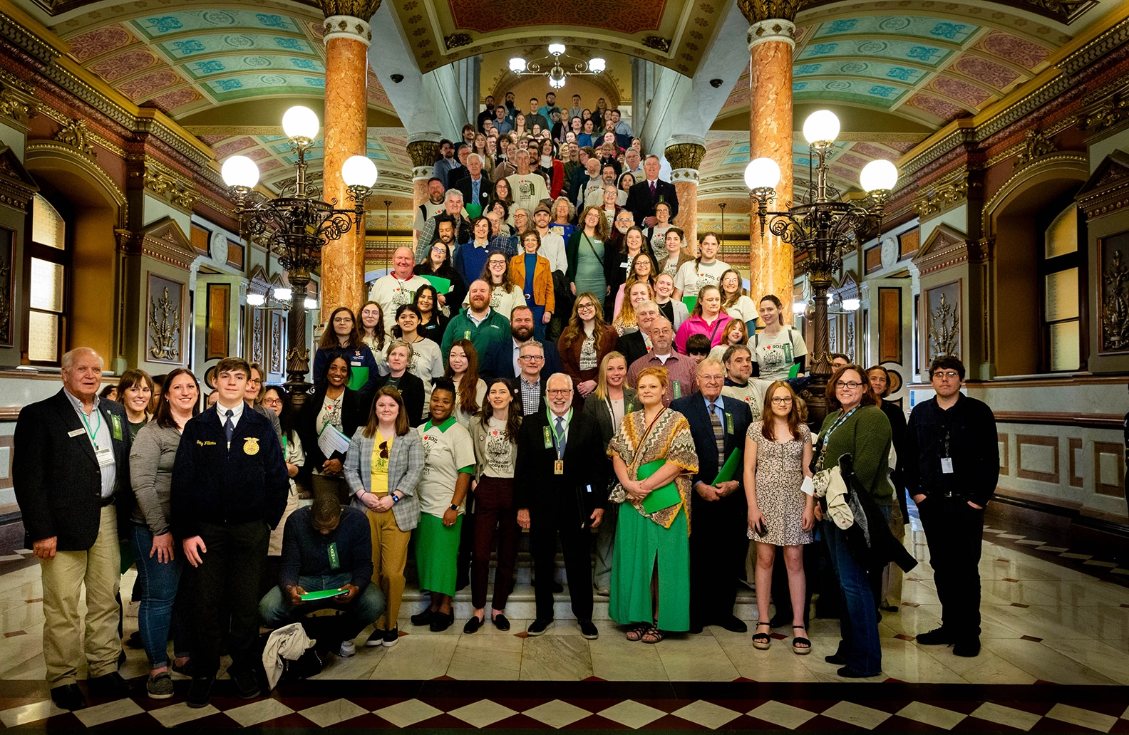 A group of food and farm advocates pose in a legislative building, surrounded by art nouveau architectural details.