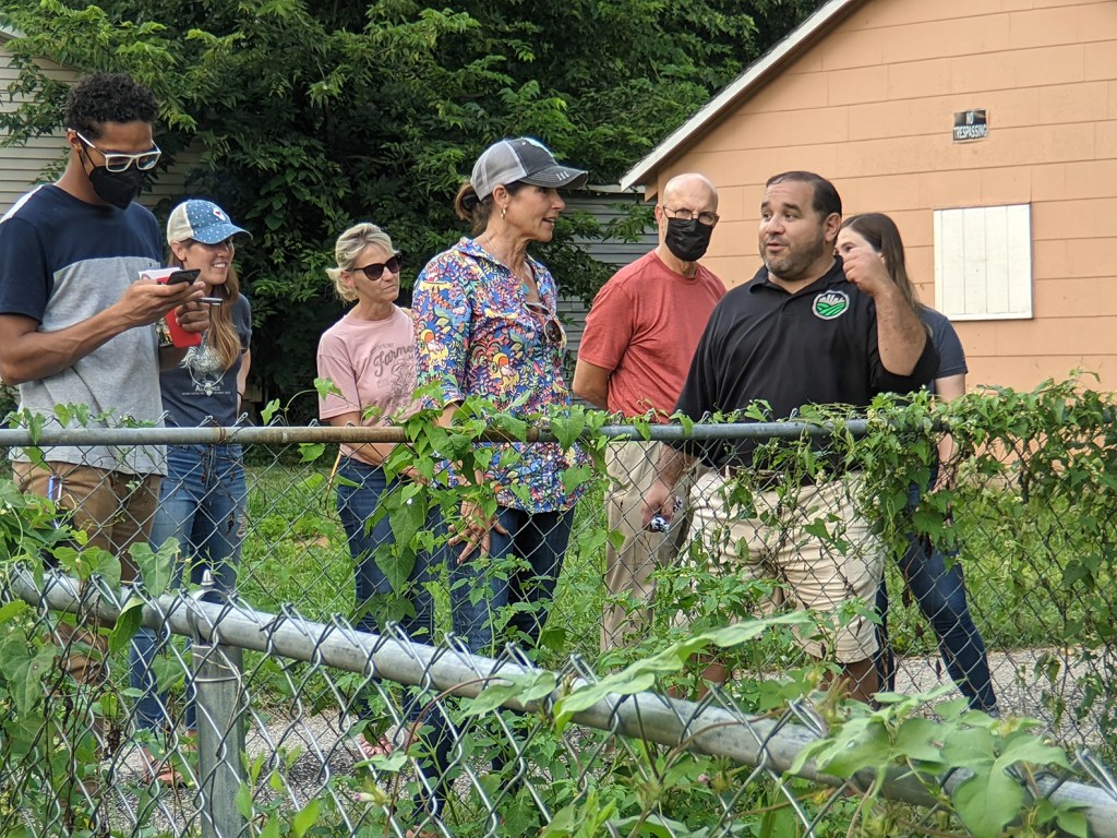 A group of farming advocates gather near a fence covered in greenery.