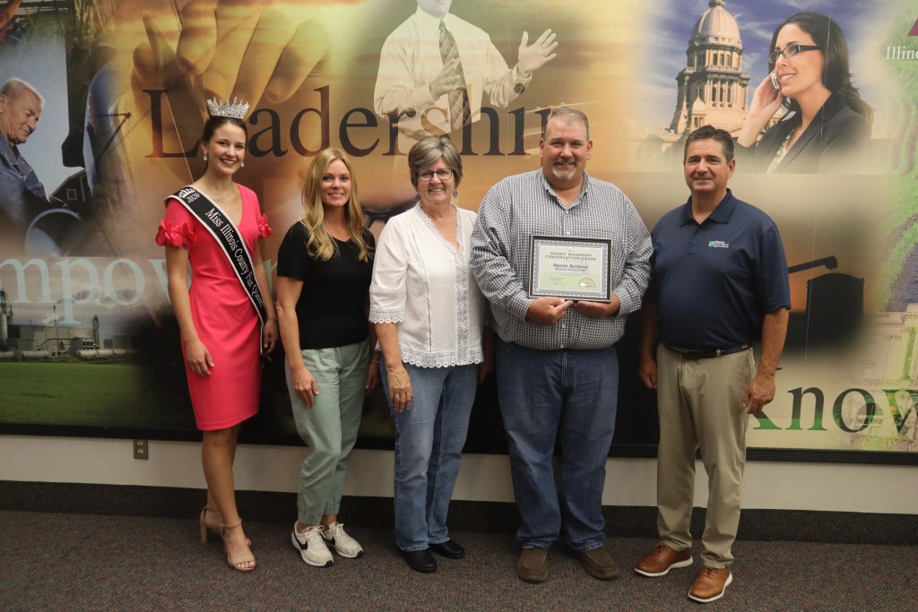 A group of five people posing for a photo, featuring a woman wearing a crown and sash titled 'Miss Illinois County.' They are standing in front of a colorful backdrop with words related to leadership and empowerment. One man is holding a certificate.