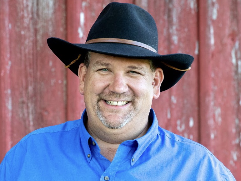 A smiling man wearing a black cowboy hat and a blue shirt, standing in front of a rustic red barn.