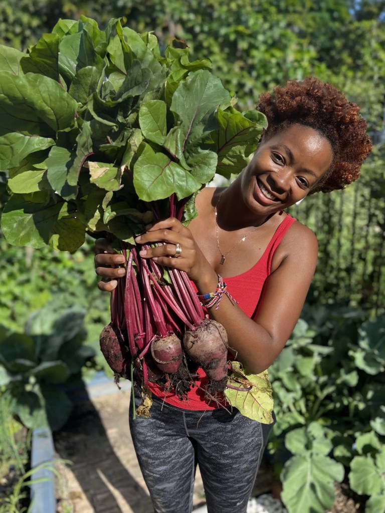 A woman smiling while holding a large bunch of freshly harvested beets with green leaves in a garden setting.