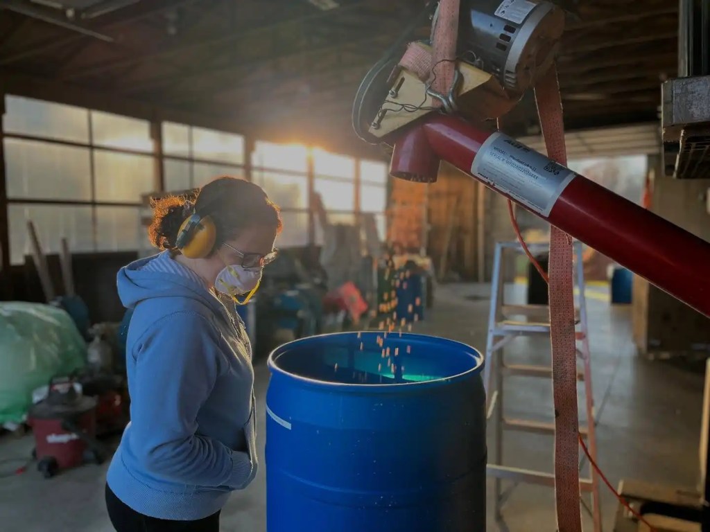 A woman wearing hearing protection and a mask observes as debris or particles fall into a blue barrel, with sunlight streaming through windows in a workshop setting.